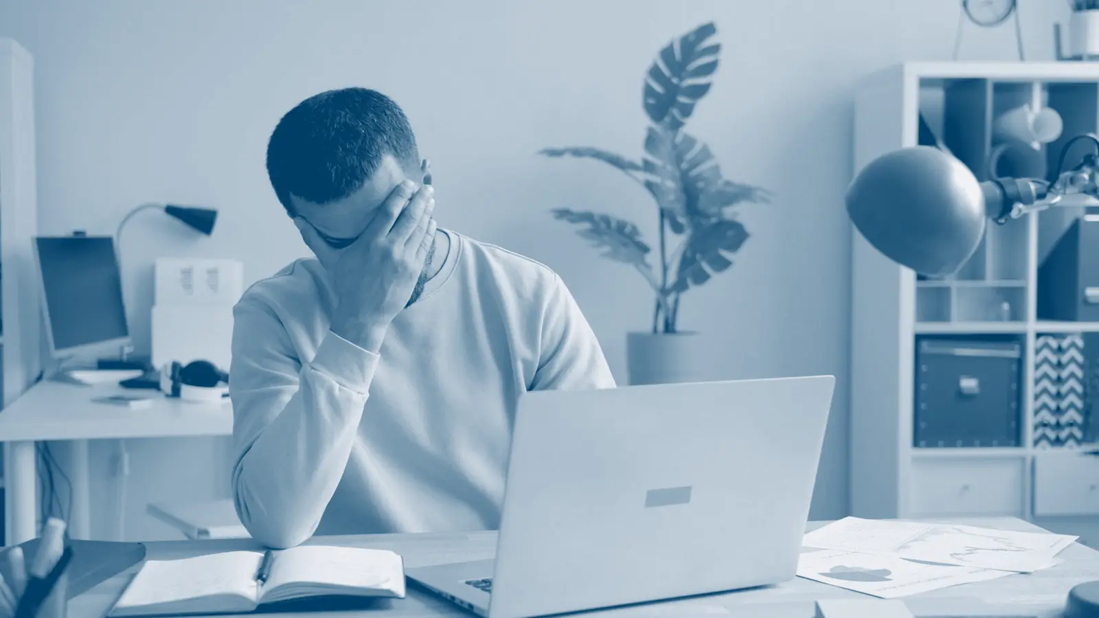 A student sitting at a laptop late at night, rubbing their face with both hands.