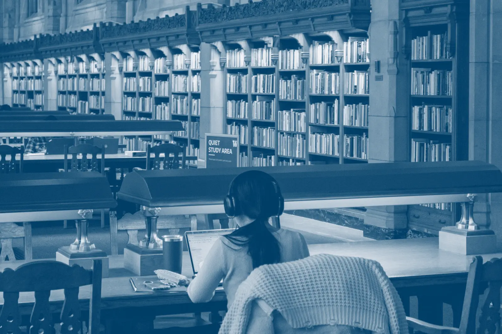 A student working at a library desk surrounded by open books.