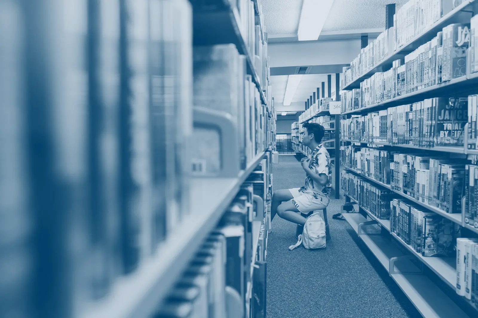 A student sitting on the floor between library shelves, reading.